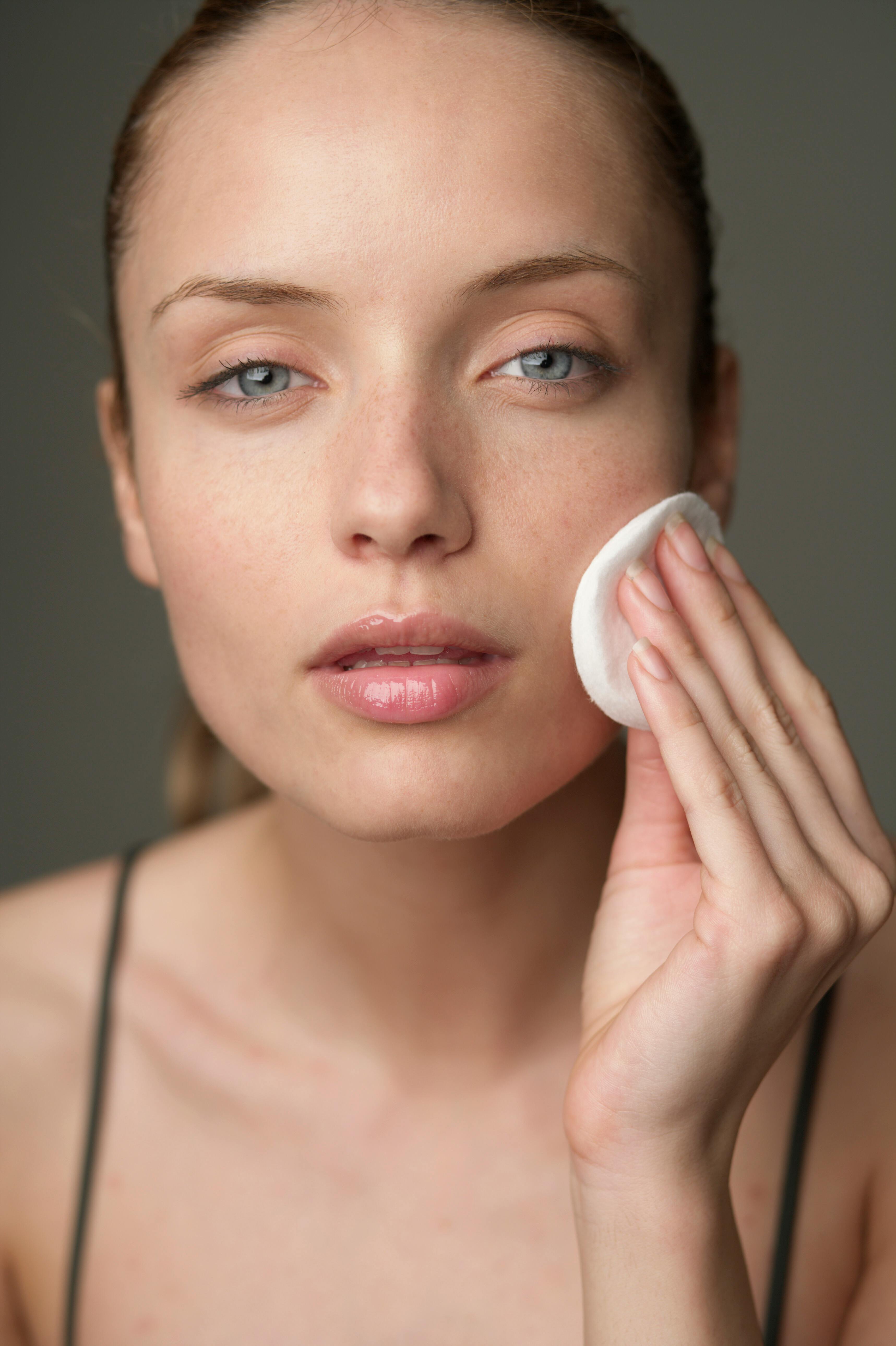 Woman applying cream to her face with a cotton pad against a neutral background